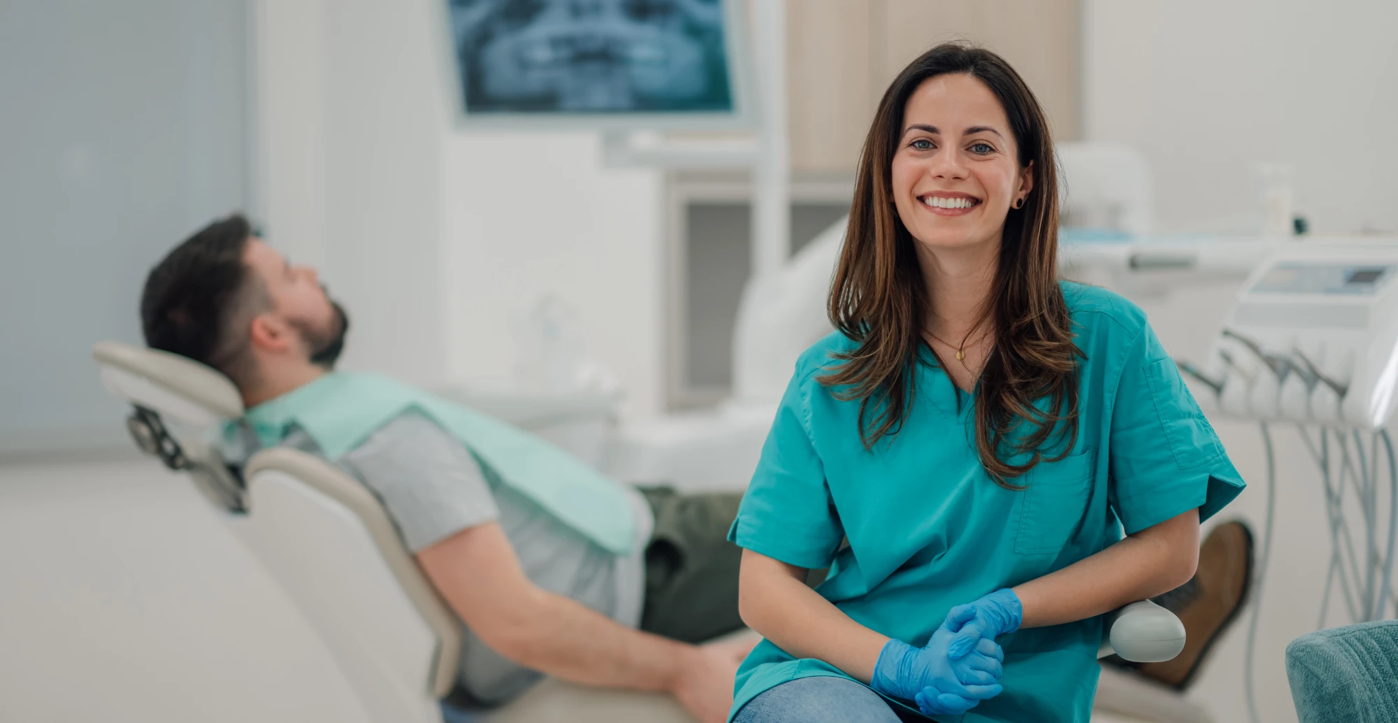 Dental professional in teal uniform smiling while working with male patient in clinic chair